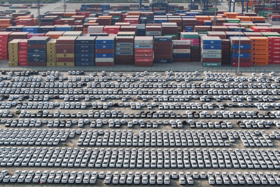 Aerial view of numerous white cars parked at a port, next to stacks of shipping containers.