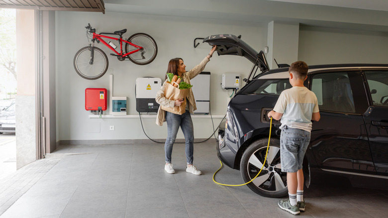 A woman carrying groceries shuts the trunk of her EV while a boy plugs in a charger