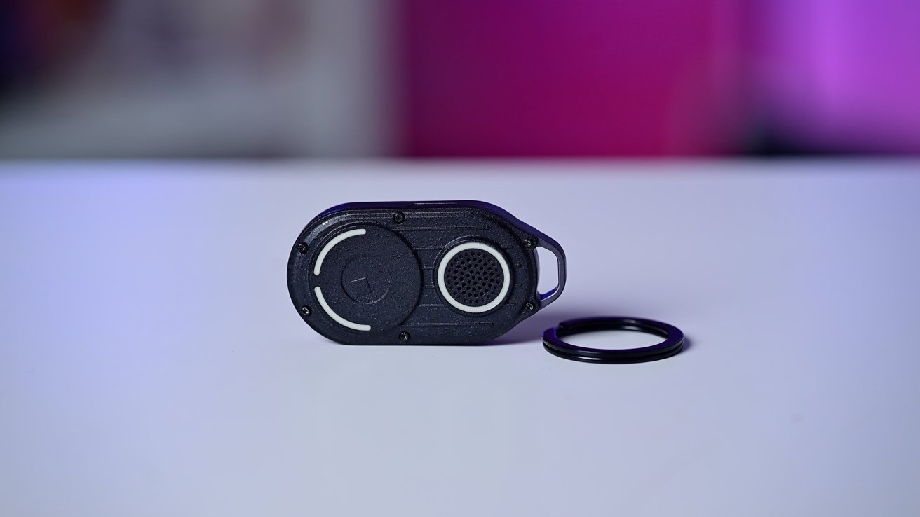 A small oval device with circular patterns and a black ring on a white surface, set against a blurred pink and purple background.