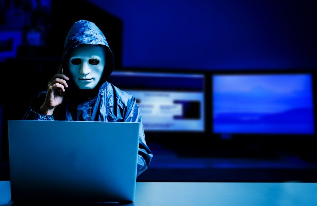 Anonymous computer hacker in white mask and hoodie. Obscured dark face using laptop computer for cyber attack and calling on cellphone, Data thief, internet attack, darknet and cyber security concept.; Shutterstock ID 1454343419; purchase_order: -; job: -; client: -; other: -