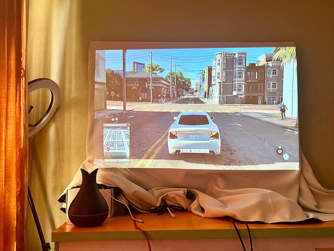 Gameplay scene projected onto white cloth showing white car on city street with mini map