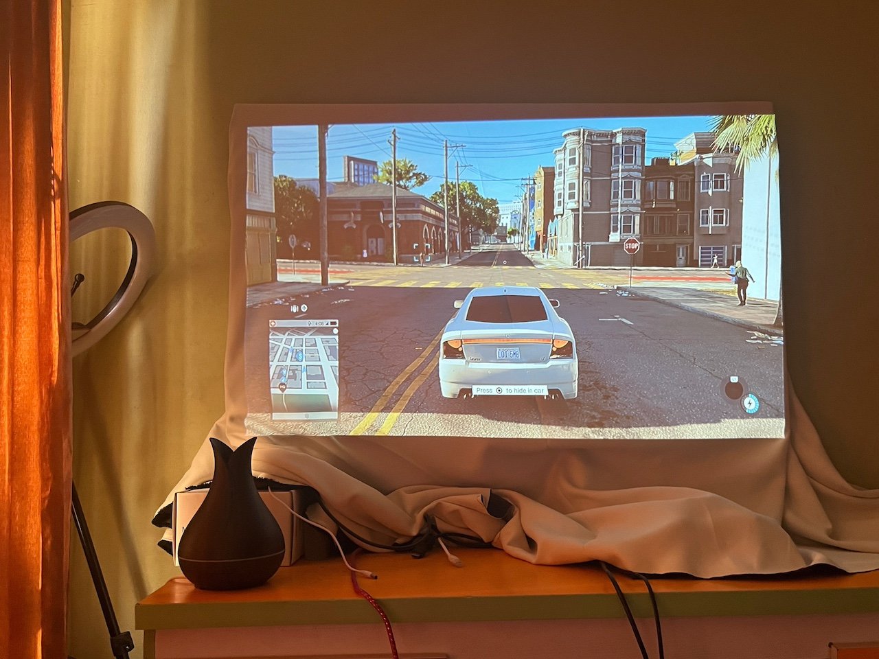 Gameplay scene projected onto white cloth showing white car on city street with mini map