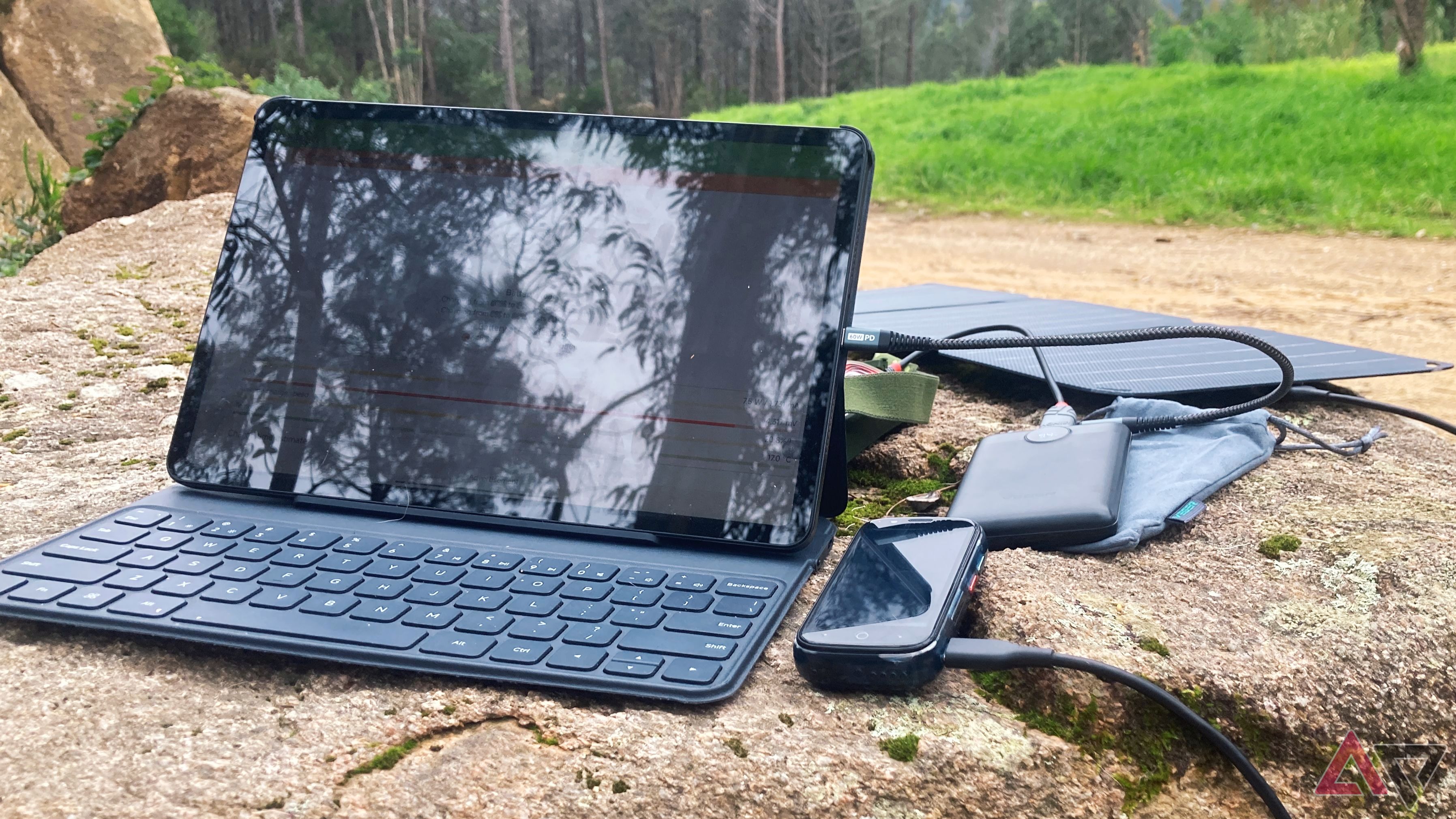 A tablet and keyboard case, smartphone, power bank, solar panel, and assortment of cables sitting on a rock outside in a nature setting.