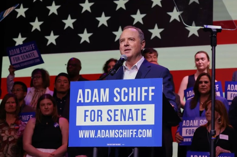 A man at a podium with "ADAM SCHIFF FOR SENATE" sign, in front of an American flag, surrounded by supporters.