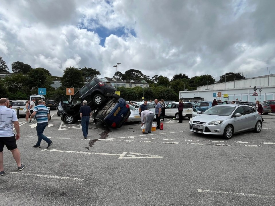 Two cars overturned in a parking lot with people standing around.