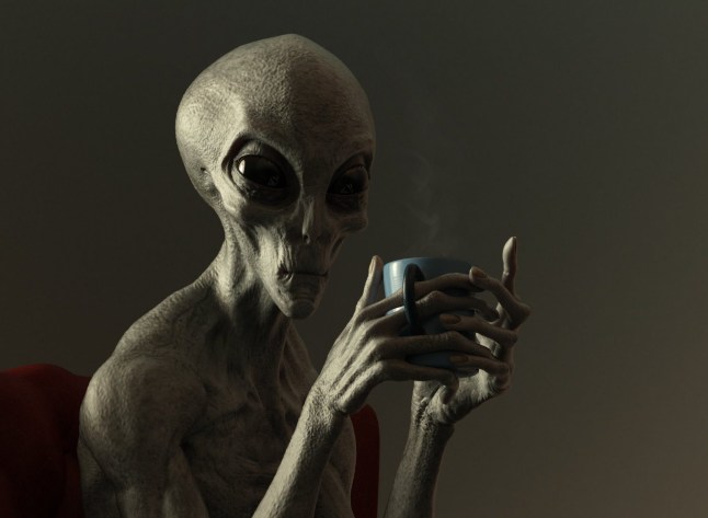 An alien sits on a red leather chair, drinking a hot beverage from a mug. He is making eye contact with the camera. The overall scene is starkly lit in low, subdued light.