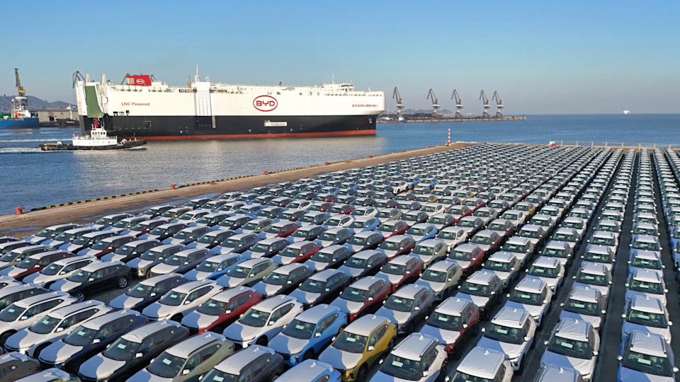 A fleet of BYD cars sit at port with a large BYD carrier in the water.
